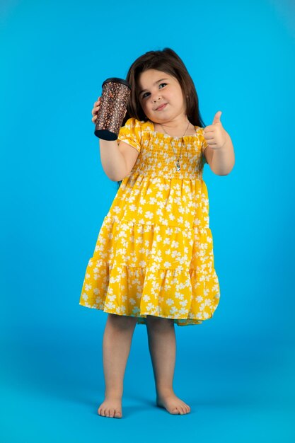 Kleines schönes lächelndes Mädchen mit einem lustigen Gesicht in einem gestreiften gelben Kleid, das auf einem blauen Hintergrund im Studio posiert