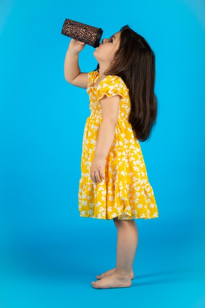 Kleines schönes lächelndes Mädchen mit einem lustigen Gesicht in einem gestreiften gelben Kleid, das auf einem blauen Hintergrund im Studio posiert