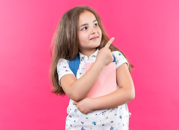 Kleines schönes Mädchen mit langen Haaren mit Rucksack, das ein Notizbuch hält, das mit einem Lächeln auf dem Gesicht aufschaut und mit dem Zeigefinger auf die Seite zeigt, die über einer rosa Wand steht?