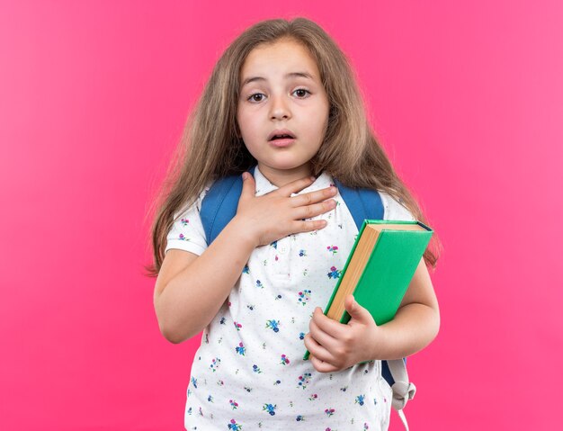 Kleines schönes Mädchen mit langen Haaren mit Rucksack, das ein Notizbuch hält, erstaunt und überrascht, die Hand auf ihrer Brust zu halten, die auf Rosa steht