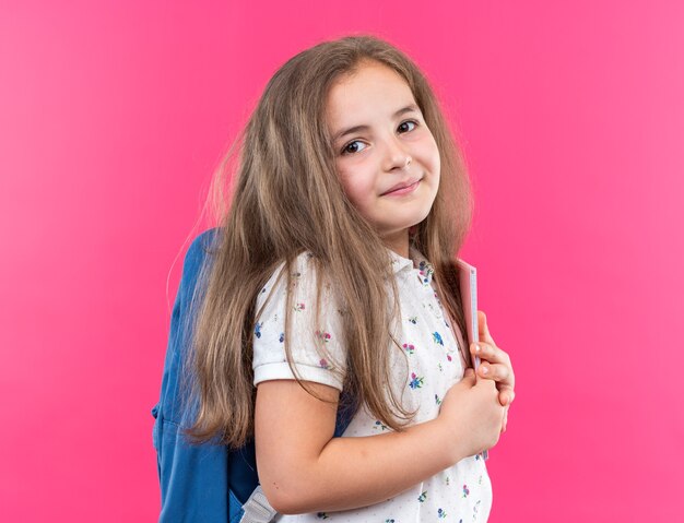 Kleines schönes Mädchen mit langen Haaren mit Rucksack, das ein Notizbuch hält und mit einem Lächeln auf einem glücklichen Gesicht über einer rosa Wand steht