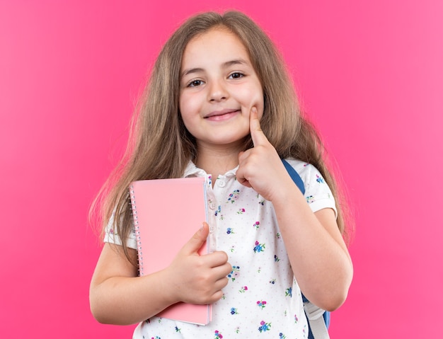Kleines schönes Mädchen mit langen Haaren mit Rucksack, das Notizbuch hält und nach vorne schaut, fröhlich lächelt und den Finger auf ihre Wange hält, die über rosa Wand steht