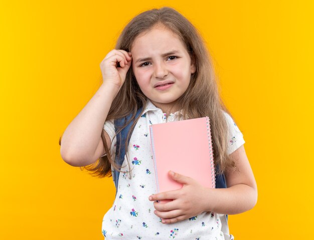 Kleines schönes Mädchen mit langen Haaren mit Rucksack, das Notizbuch verwirrt und sehr ängstlich auf Orange hält