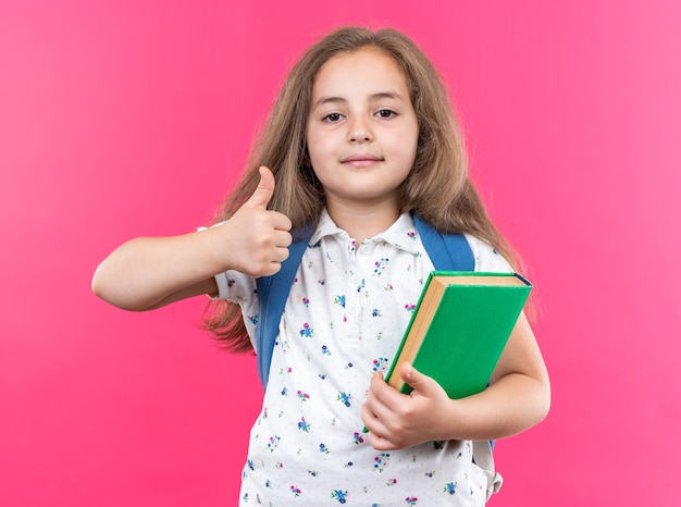 Kleines schönes Mädchen mit langen Haaren mit Rucksack mit Notebook, das nach vorne schaut und selbstbewusst lächelt und Daumen nach oben über rosa Wand steht