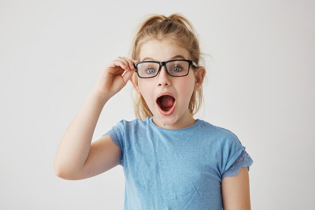 Kleines schönes Mädchen mit strahlend blauen Augen und blonden Haaren, die mit geöffnetem Mund aufwerfen und ihre Brille mit der Hand halten, die super überrascht aussieht.