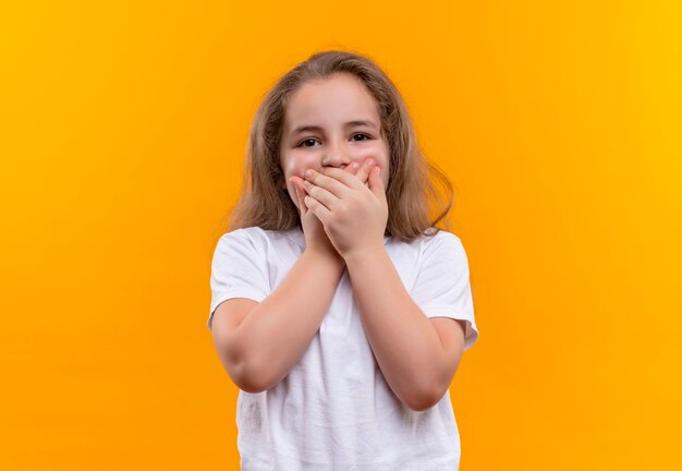 kleines Schulmädchen, das den weißen T-Shirt bedeckten Mund mit beiden Händen auf isolierter orange Wand trägt