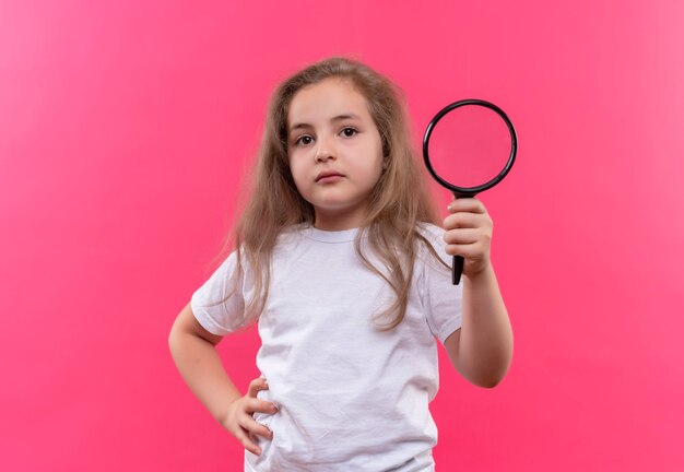 kleines Schulmädchen, das weißes T-Shirt hält, das Lupe auf isolierter rosa Wand hält