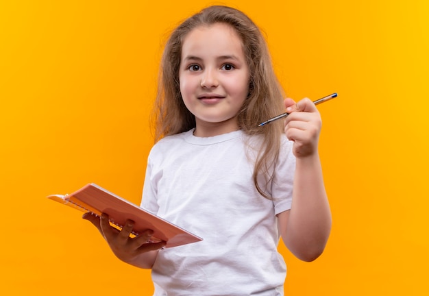 kleines Schulmädchen, das weißes T-Shirt hält, das Notizbuch und Stift auf isolierter orange Wand hält