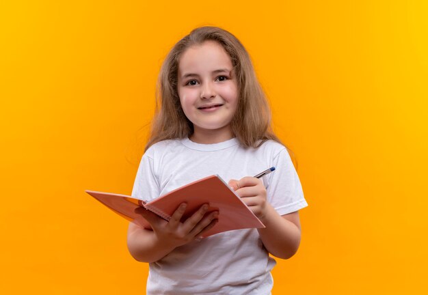 kleines Schulmädchen, das weißes T-Shirt hält, das Notizbuch und Stift auf isolierter orange Wand hält