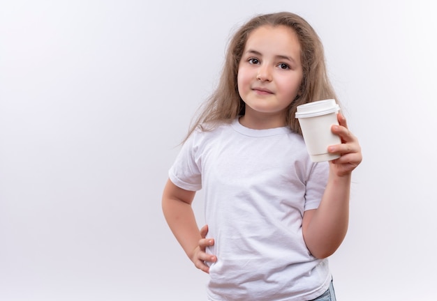 kleines Schulmädchen, das weißes T-Shirt hält, das Tasse Kaffee hält, legte ihre Hand auf Hüfte auf isolierte weiße Wand
