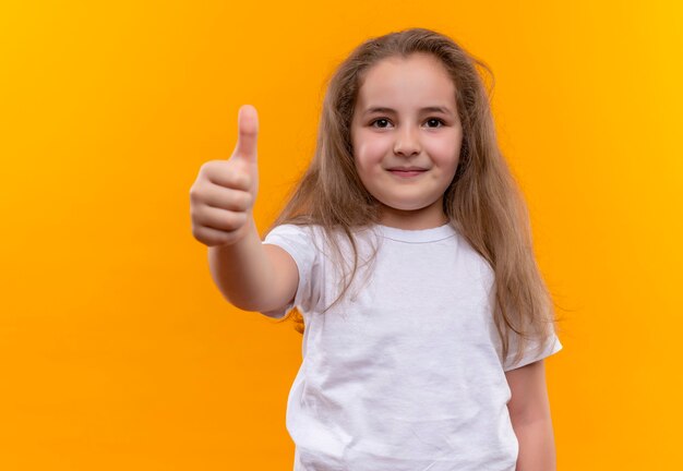 kleines Schulmädchen, das weißes T-Shirt ihren Daumen oben auf isolierter orange Wand trägt