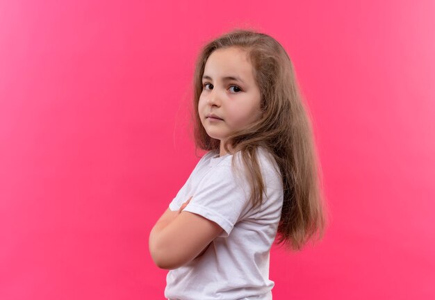 kleines Schulmädchen, das weißes T-Shirt trägt, das Hände auf isolierter rosa Wand kreuzt