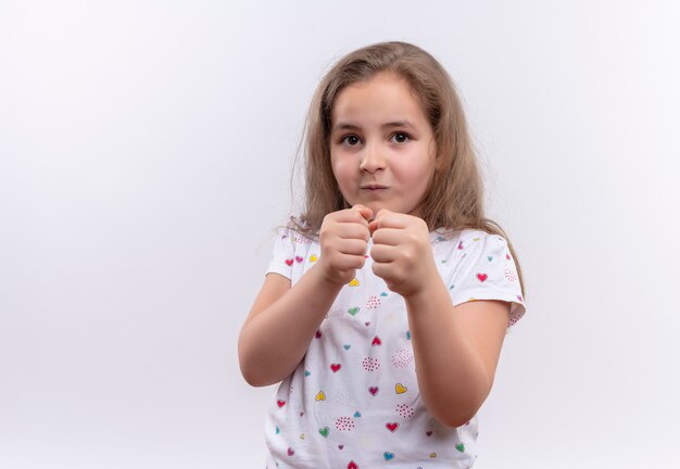 kleines Schulmädchen, das weißes T-Shirt trägt, das in der Kampfhaltung auf isolierter weißer Wand steht