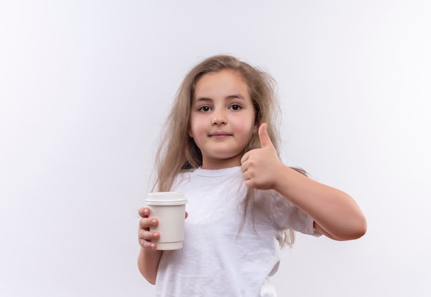 kleines Schulmädchen, das weißes T-Shirt trägt, das Tasse Kaffee ihren Daumen oben auf isolierter weißer Wand hält