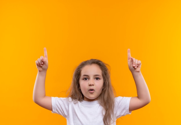 kleines Schulmädchen, das weißes T-Shirt trägt, legte ihren Finger auf isolierte orange Wand