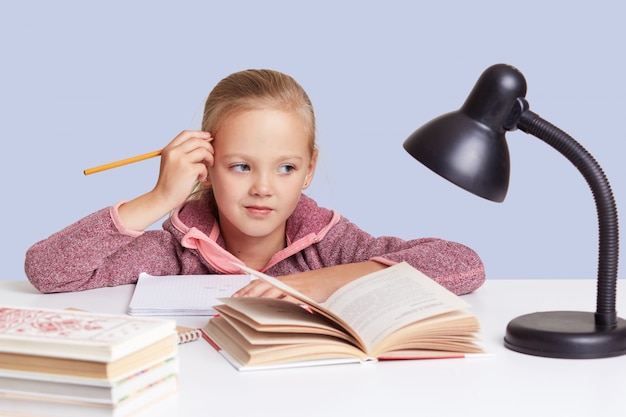 Kleines schulmädchen hält die hand in der nähe des kopfes, schaut nachdenklich aus, denkt über hausaufgaben nach, benutzt eine leselampe. kinder-, bildungs- und schulkonzept.