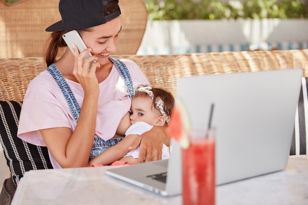 Kleines süßes Baby füttert von der Brust der Mutter. Die fröhliche junge Mutter spricht mit einer Freundin per Handy und kümmert sich um ihr Kind.