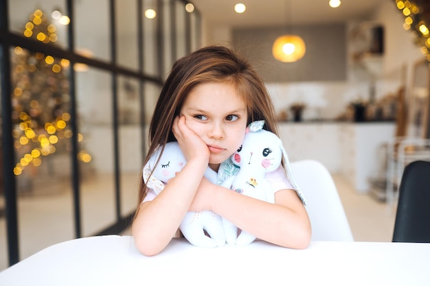 Kleines trauriges Mädchen, das Lieblingsspielzeug umarmt, während es am Tisch sitzt, glückliche Kindheit