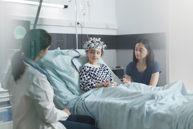 Klinischer pädiatrischer Experte, der die EEG-Scan-Ergebnisse eines kranken Mädchens überprüft, während er sich im Beratungsraum der Kinderstation befindet. Ärztin und ängstliche Mutter diskutieren über Behandlung und Erholungszeit.