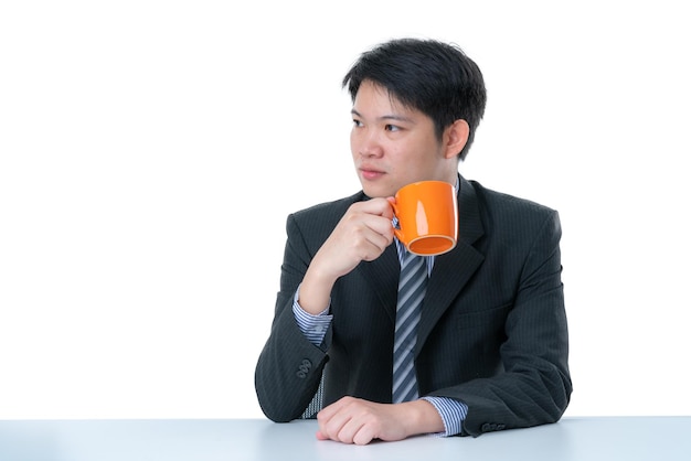 Kluger asiatischer geschäftsmann im anzug sitzt am schreibtisch und trinkt heißen kaffee auf weißem hintergrund