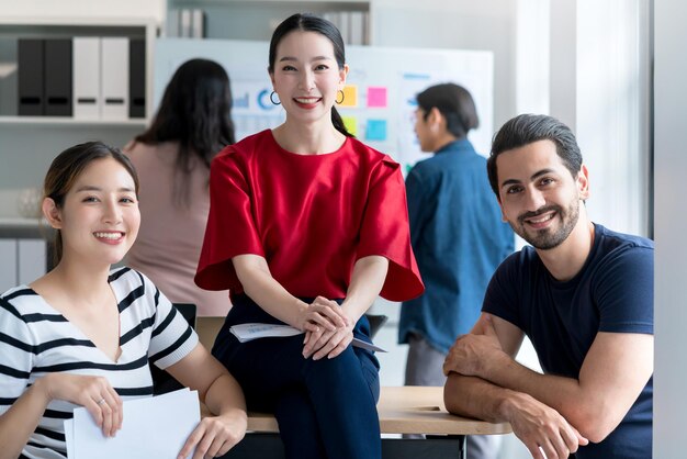 Kluger asiatischer Startup-Kleinunternehmer, der selbstbewusstes Porträt mit Freunden, Partner, Gelegenheitstreffen, Brainstorming mit Whiteboard und Geschäftsfinanzierungspapier-Diagrammanalyse im Bürohintergrund steht