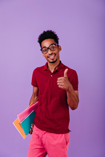 Kluger schwarzer Student, der mit Daumen oben aufwirft. Innenfoto des erfreuten afrikanischen Kerls mit Büchern, die Spaß nach Prüfungen haben.