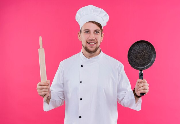 Koch, der Kameralächeln auf Gesicht mit Nudelholz und Pfanne betrachtet, die über rosa Wand stehen