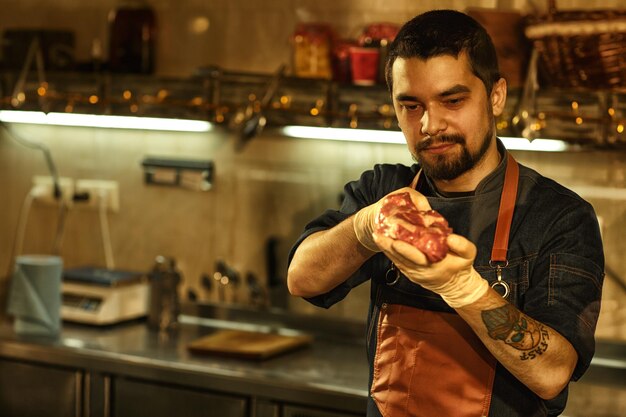 Koch, der sich Steak ansieht und die Qualität des Fleisches überprüft Schöner Mann mit Tattoo auf seinem Arm mit Schürze und weißen Handschuhen Hintergrund der professionellen Restaurantküche mit Geschirr