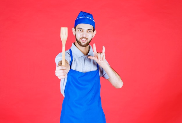 Koch in blauer Schürze, der einen Holzspatel hält und Genusszeichen zeigt.