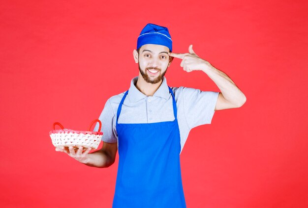 Koch in blauer Schürze, die einen mit rotem Tuch bedeckten Brotkorb hält und denkt.