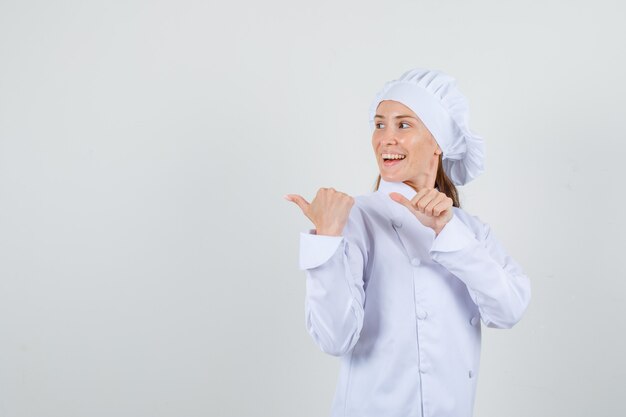 Köchin, die Daumen in weißer Uniform zur Seite zeigt und fröhlich aussieht.