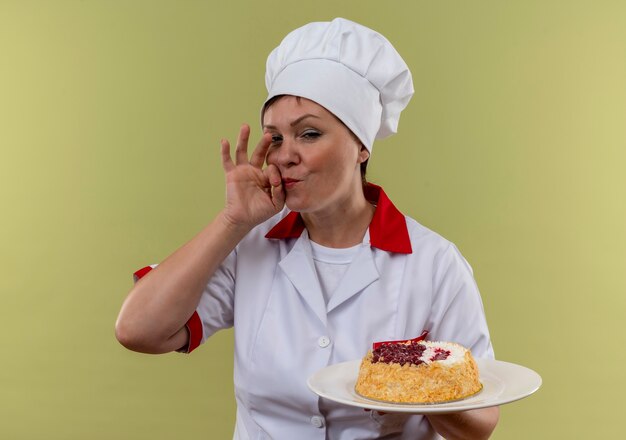 Köchin mittleren Alters in der Kochuniform, die Kuchen auf Teller hält und köstliche Geste auf isolierter grüner Wand zeigt