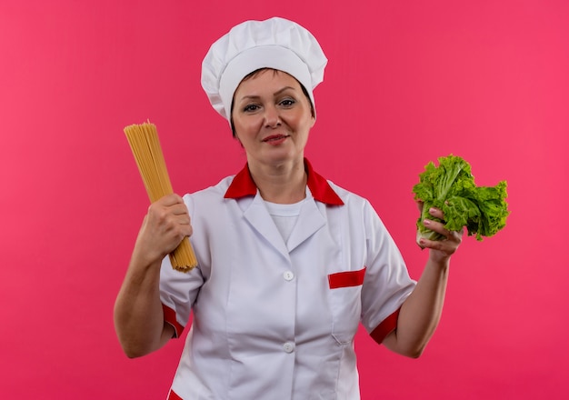 Köchin mittleren Alters in der Kochuniform, die Salat und Spaghetti auf isolierter rosa Wand hält