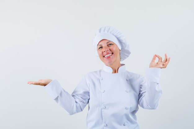 Köchin zeigt Siegergeste, während sie Handfläche in weißer Uniform beiseite spreizt und erfreut aussieht.