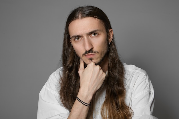 Körpersprache. Porträt eines hübschen unrasierten jungen europäischen Mannes mit Schnurrbart und langer loser Frisur, die das Kinn berührt, über eine Idee, ein Problem oder ein Projekt nachdenkend, isoliert posierend