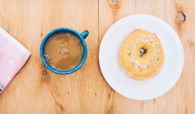 Köstlicher Donut auf Teller; Kaffeetasse und Serviette auf strukturiertem Hintergrund aus Holz