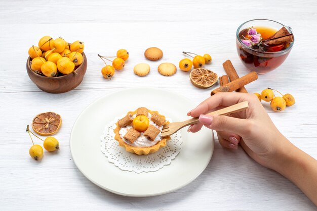 köstlicher kleiner Kuchen, der von Frau zusammen mit gelben Kirschen-Zimt-Coookies und Tee auf leichtem Schreibtisch, Kekskuchen-Keks süß gegessen wird