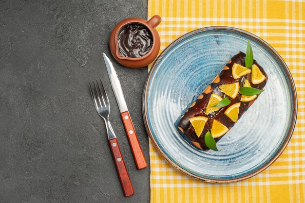 Köstlicher Kuchen, dekoriert mit Orange und Schokolade, serviert mit Gabel und Messer auf dunklem Tisch