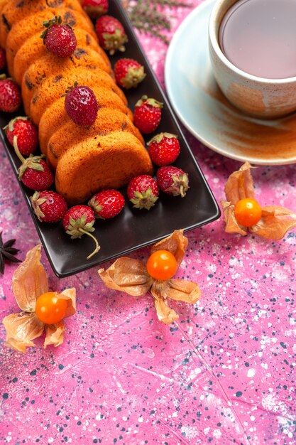 Köstlicher Kuchen der halben Draufsicht in der schwarzen Kuchenform mit frischen roten Erdbeeren und einer Tasse Tee auf rosa Schreibtisch.