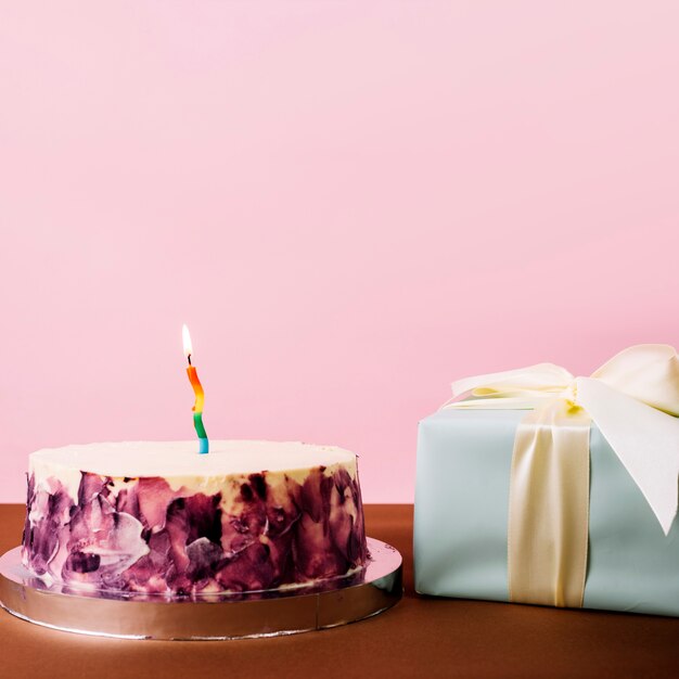 Köstlicher Kuchen mit belichteter Kerze und eingewickelter Geschenkbox gegen rosa Hintergrund