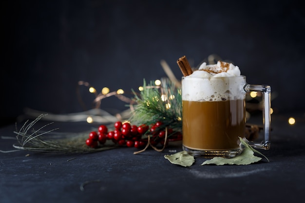 Köstlicher Weihnachtskaffee mit Zimt und Schaum, neben Hollies auf dunkler Oberfläche