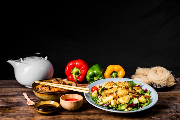 Köstliches thailändisches Essen mit Sojasauce; Teekanne und Paprika auf Schreibtisch vor schwarzem Hintergrund