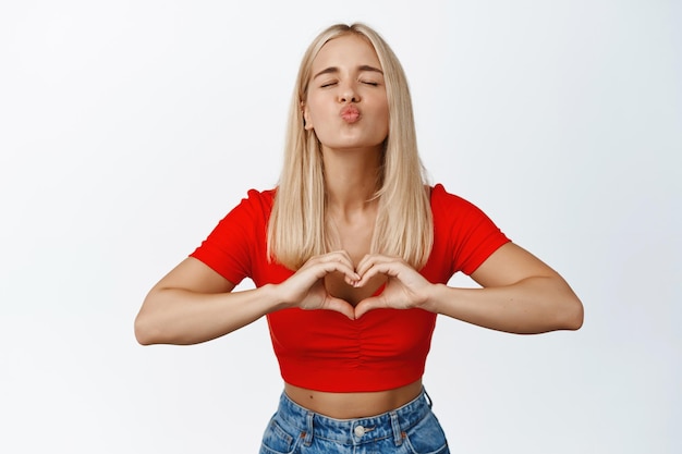 Kokettes blondes Mädchen zeigt küssendes Gesicht und Herzzeichen, die über weißem Hintergrund in stilvoller Sommerkleidung stehen