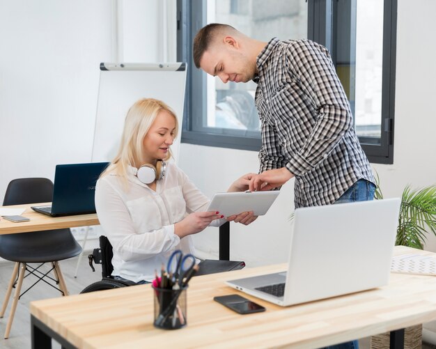 Kollege zeigt Frau im Rollstuhl etwas auf Tablette bei der Arbeit