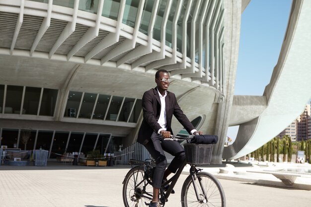 Ökologisch bewusster junger schwarzer europäischer Geschäftsmann, der schwarzen Anzug und Sonnenbrille trägt, die während der Mittagspause am sonnigen Tag zum Café auf Retro-Fahrrad radeln
