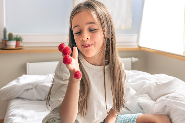 Komisches kleines Mädchen mit Himbeeren an den Fingern im Bett am Morgen