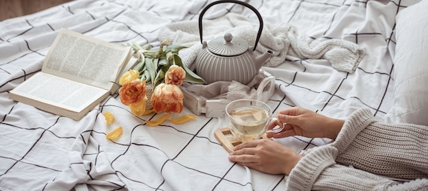 Komposition mit einer Tasse Tee, einer Teekanne, einem Strauß Tulpen und einem Buch im Bett