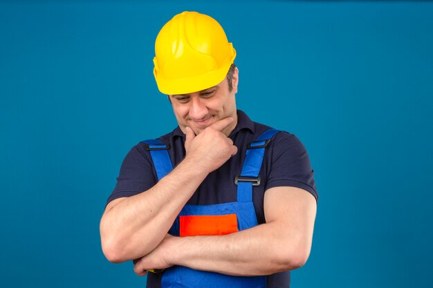 Konstrukteur mittleren Alters, der Konstruktionsuniform und Sicherheitshelm trägt, der Kinn berührt und über isolierte blaue Wand denkt