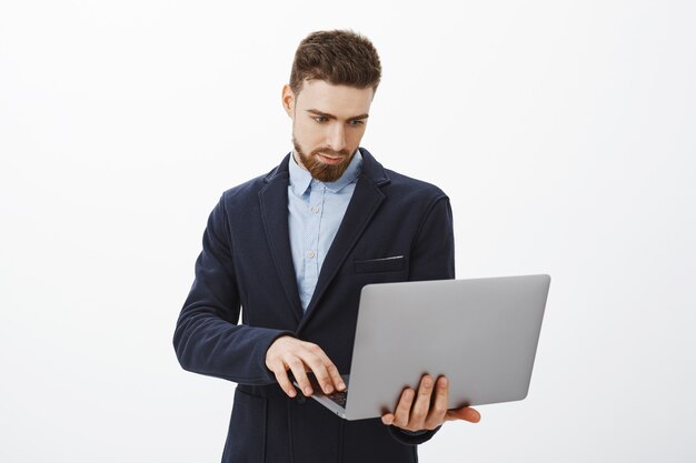 Konzentration auf das Geschäft. Porträt des intelligenten und ehrgeizigen konzentrierten gutaussehenden jungen männlichen Unternehmers mit Bart und blauen Augen, die Laptop in der Hand halten, die Prüfplan mit entschlossenem Blick durchsucht