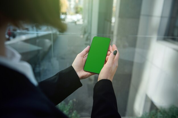 Konzentrieren sie sich auf ein telefon mit einem grünen chroma-key, um anzeigen in die hände einer nicht erkennbaren frau einzufügen, die im fenster eines hochhauses mit blick auf einen städtischen hintergrund steht. platz kopieren. nahaufnahme